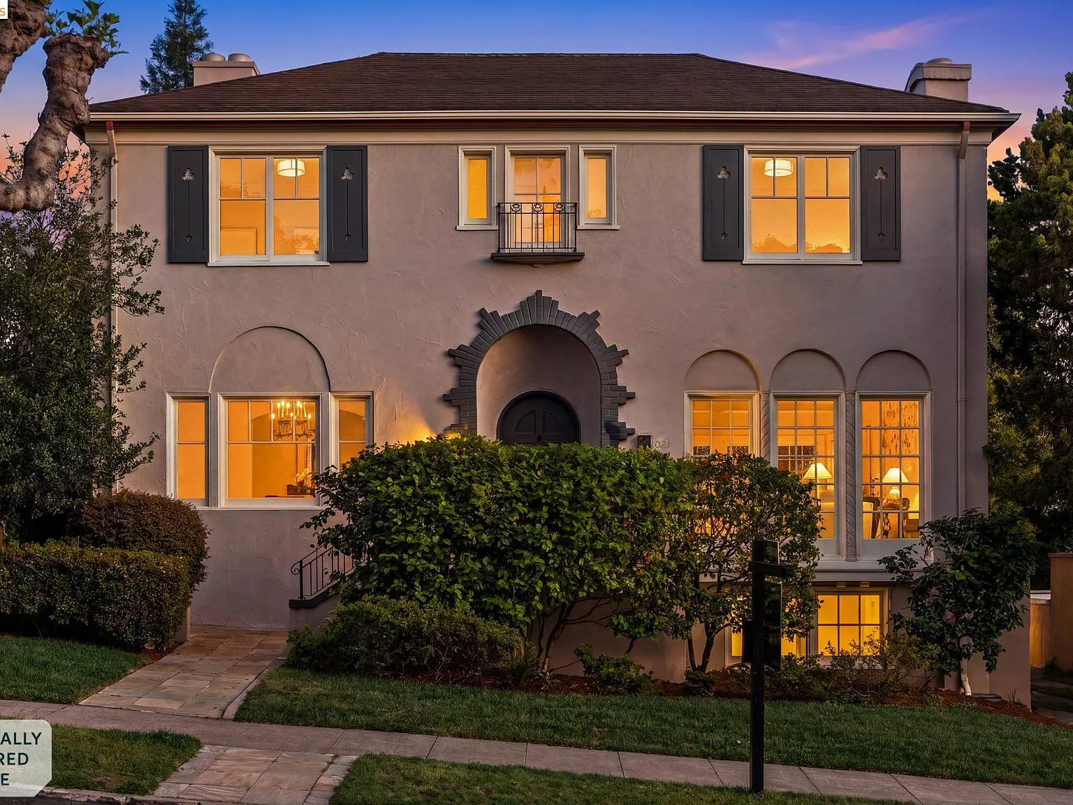 A stucco Spanish Colonial home down the street from 635 Longridge with arched doorway and tall shuttered windows, lit warmly from within at dusk